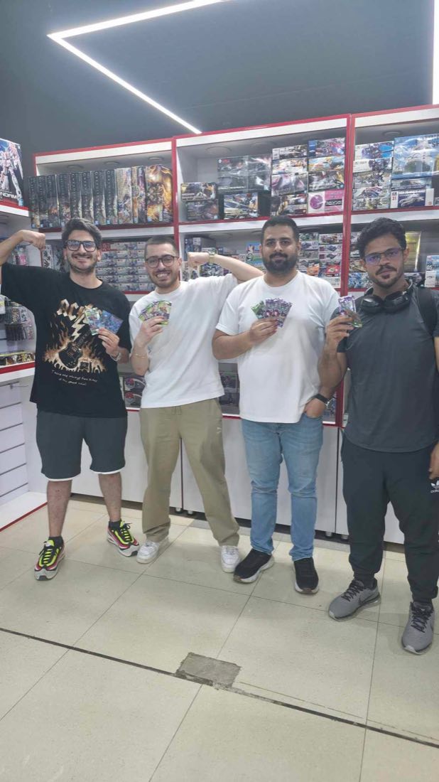 Four players holding booster packs in front of the model kit shelves